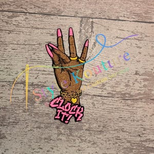 May include: Embroidered patch featuring a brown hand with pink fingernails and gold rings and bracelets. The patch has the text "CLOCK IT!" in pink with a black outline. The patch is on a wooden surface.