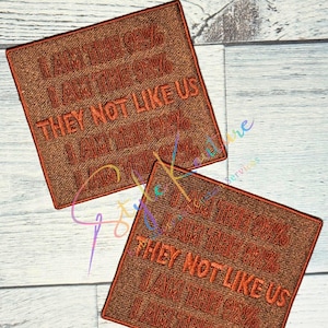 May include: Two brown embroidered patches with the text "I AM THE 99%" and "THEY NOT LIKE US" in orange thread. The patches are square and have a textured appearance, with a contrasting orange border.