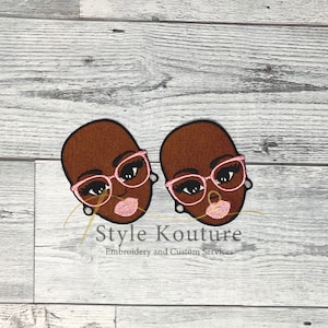 May include: Two embroidered patches of brown-skinned women wearing pink glasses and pink lipstick. The patches are on a light wood background.