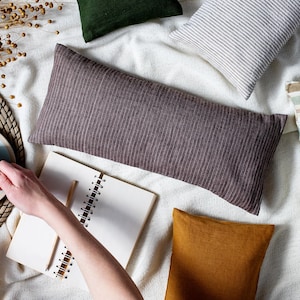 May include: A flat lay of a white blanket with various pillows, including a brown striped lumbar pillow, a white and gray striped pillow, a green pillow, and a mustard yellow pillow. A notebook and a cup of coffee are also on the blanket.