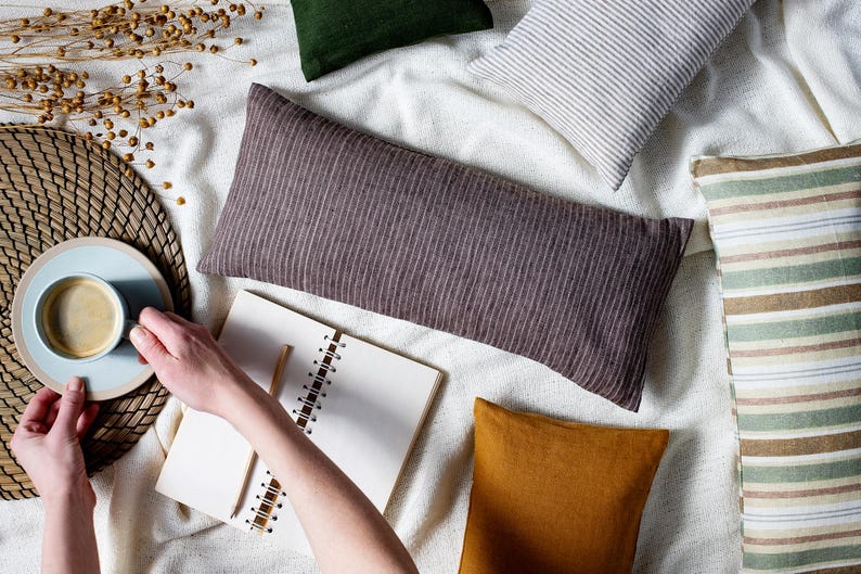 May include: A flat lay of a white blanket with various pillows, including a brown striped lumbar pillow, a white and gray striped pillow, a green pillow, and a mustard yellow pillow. A notebook and a cup of coffee are also on the blanket.