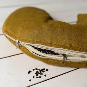 May include: A mustard yellow crescent-shaped pillow with a zipper opening, revealing buckwheat hulls inside. Some buckwheat hulls are scattered on a white wooden surface.
