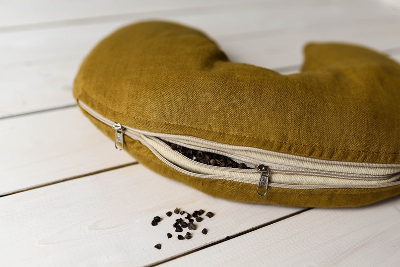May include: A mustard yellow crescent-shaped pillow with a zipper opening, revealing buckwheat hulls inside. Some buckwheat hulls are scattered on a white wooden surface.