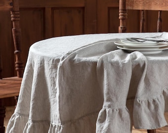 May include: A round, white linen tablecloth with thin black stripes and a ruffled edge. The tablecloth is draped over a wooden table with a chair visible in the background.