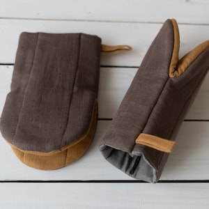 Può includere: Due guanti da forno marroni con bordino beige. Un guanto è in stile presina e l'altro è un guanto completo per la mano.