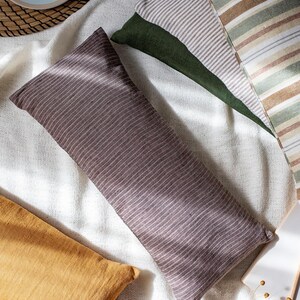 May include: Three decorative pillows with striped patterns in shades of brown, beige, green, and white. The pillows are arranged on a white linen fabric background.