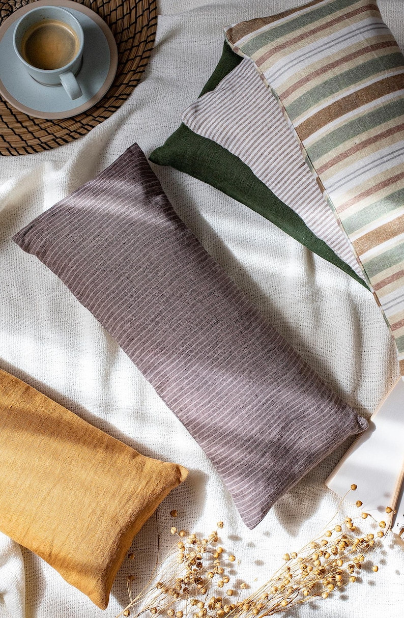 May include: Three decorative pillows with striped patterns in shades of brown, beige, green, and white. The pillows are arranged on a white linen fabric background.