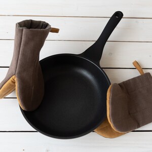 May include: Two brown oven mitts with tan accents surround a black cast iron skillet on a white wooden surface.