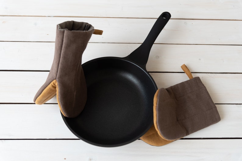 May include: Two brown oven mitts with tan accents surround a black cast iron skillet on a white wooden surface.