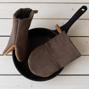 May include: Two brown oven mitts with tan trim, one is folded and placed inside a black cast iron skillet with a black handle.