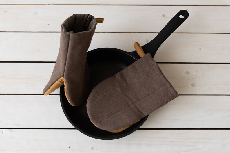May include: Two brown oven mitts with tan trim, one is folded and placed inside a black cast iron skillet with a black handle.