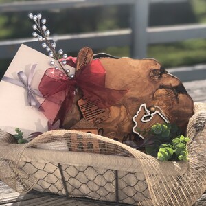 Puede incluir: Una tabla de servir de madera r&uacute;stica con un borde natural, un peque&ntilde;o letrero de madera con el texto "Gracias", un regalo envuelto y una rama de vegetaci&oacute;n en una cesta de alambre forrada de arpillera.
