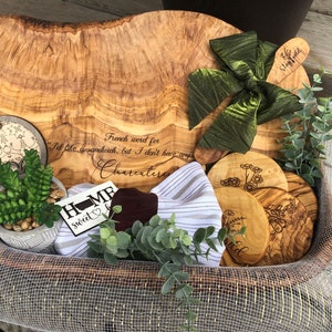 Puede incluir: Una cesta de mimbre llena de art&iacute;culos de madera de olivo, incluyendo una tabla de servir grande, una tabla de cortar m&aacute;s peque&ntilde;a y varios posavasos. La tabla de servir tiene una cita grabada: "Palabra francesa para Me gustar&iacute;a un s&aacute;ndwich, pero no tengo charcuter&iacute;a". La cesta tambi&eacute;n contiene una peque&ntilde;a suculenta en maceta, un pa&ntilde;o de cocina a rayas verde y blanco y un letrero de madera que dice "Home Sweet Home".