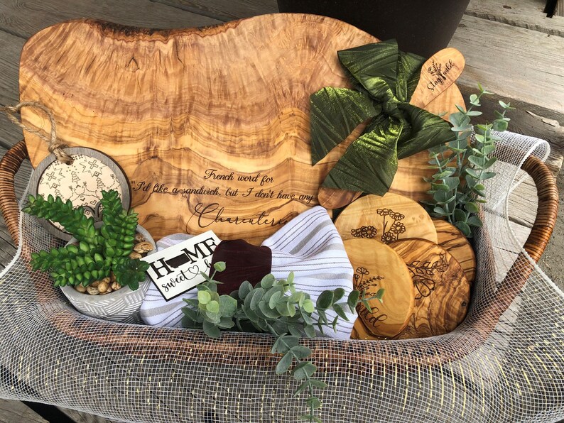 Puede incluir: Una cesta de mimbre llena de art&iacute;culos de madera de olivo, incluyendo una tabla de servir grande, una tabla de cortar m&aacute;s peque&ntilde;a y varios posavasos. La tabla de servir tiene una cita grabada: "Palabra francesa para Me gustar&iacute;a un s&aacute;ndwich, pero no tengo charcuter&iacute;a". La cesta tambi&eacute;n contiene una peque&ntilde;a suculenta en maceta, un pa&ntilde;o de cocina a rayas verde y blanco y un letrero de madera que dice "Home Sweet Home".