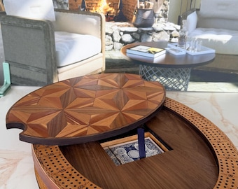May include: An oval wooden cribbage board featuring a geometric star pattern on its removable lid. The board has a dark wood center with a recessed compartment containing a deck of playing cards and a blue peg, surrounded by a track of holes.