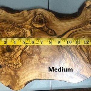 Puede incluir: Una tabla de cortar de madera de olivo de tama&ntilde;o mediano con una forma natural irregular. La tabla mide 38 cm de largo. La madera tiene un rico color marr&oacute;n y dorado con un patr&oacute;n de veta distintivo.