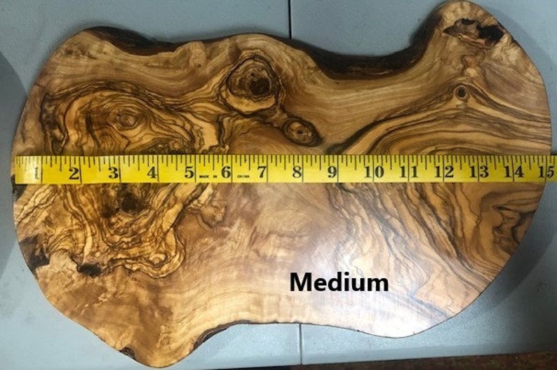 Puede incluir: Una tabla de cortar de madera de olivo de tama&ntilde;o mediano con una forma natural irregular. La tabla mide 38 cm de largo. La madera tiene un rico color marr&oacute;n y dorado con un patr&oacute;n de veta distintivo.