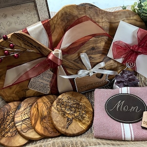 Puede incluir: Una cesta de regalo llena de art&iacute;culos de madera de olivo, que incluyen una tabla de servir grande, cuatro posavasos, una peque&ntilde;a esp&aacute;tula de madera y un peque&ntilde;o letrero de madera que dice "Pensilvania". La cesta est&aacute; forrada de arpillera y decorada con cinta roja y blanca.