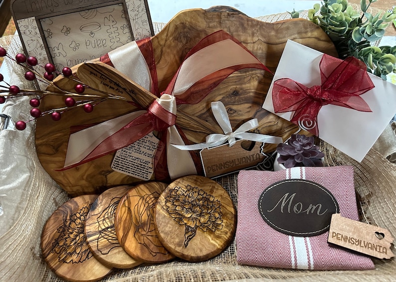 Puede incluir: Una cesta de regalo llena de art&iacute;culos de madera de olivo, que incluyen una tabla de servir grande, cuatro posavasos, una peque&ntilde;a esp&aacute;tula de madera y un peque&ntilde;o letrero de madera que dice "Pensilvania". La cesta est&aacute; forrada de arpillera y decorada con cinta roja y blanca.