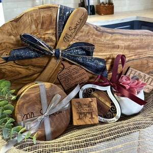 Puede incluir: Una cesta de regalo llena de art&iacute;culos de madera de olivo, que incluyen una tabla de cortar grande, dos posavasos, un peque&ntilde;o letrero de madera que dice "Home" y un adorno en forma de coraz&oacute;n. La cesta est&aacute; forrada con arpillera y decorada con una cinta azul y dorada.