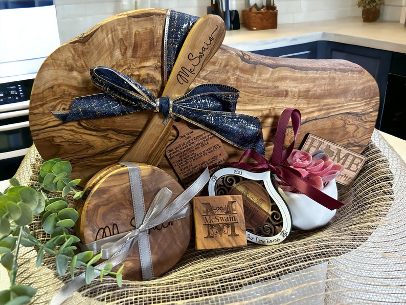 Puede incluir: Una cesta de regalo llena de art&iacute;culos de madera de olivo, que incluyen una tabla de cortar grande, dos posavasos, un peque&ntilde;o letrero de madera que dice "Home" y un adorno en forma de coraz&oacute;n. La cesta est&aacute; forrada con arpillera y decorada con una cinta azul y dorada.
