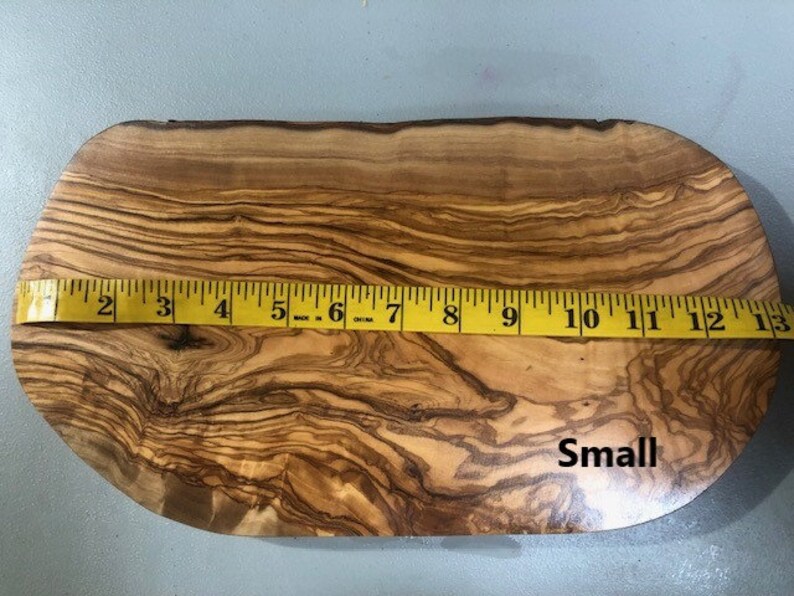 Puede incluir: Una tabla de cortar de madera de olivo peque&ntilde;a, de forma ovalada, con un patr&oacute;n de veta de madera natural. La tabla mide 13 pulgadas de largo y 10 pulgadas de ancho en su punto m&aacute;s ancho. La tabla est&aacute; etiquetada como "Small".