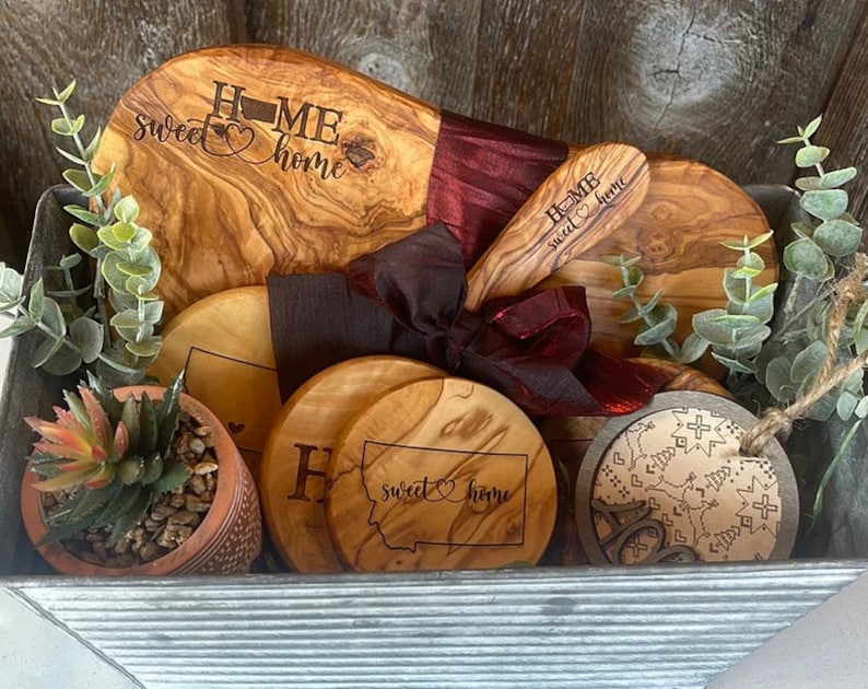 Puede incluir: Una cesta de regalo llena de piezas de servicio de madera de olivo, que incluyen una tabla de cortar grande, una tabla de servir m&aacute;s peque&ntilde;a y cuatro posavasos. Las piezas de servicio est&aacute;n grabadas con la frase "Home sweet home" y un coraz&oacute;n. La cesta tambi&eacute;n incluye una peque&ntilde;a planta suculenta en maceta y un adorno de madera decorativo.