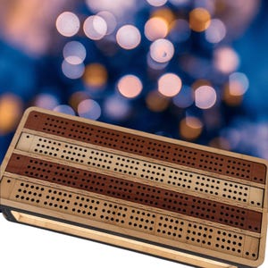 May include: A wooden cribbage board with alternating stripes of light and dark wood. The board has numerous small holes for peg placement and a dark border. The background is blurred with bokeh lights.