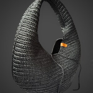 May include: A crescent-shaped hobo bag crafted from black woven material with a textured surface. The bag features a single integrated shoulder strap and a small tan leather tab detail secured with a metallic rivet near the opening.