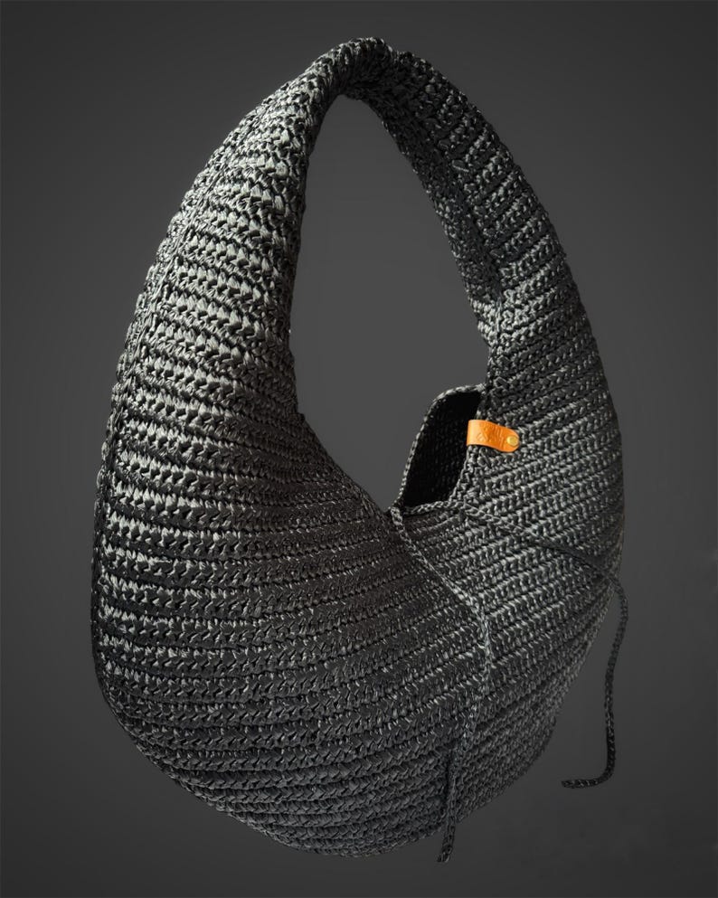 May include: A crescent-shaped hobo bag crafted from black woven material with a textured surface. The bag features a single integrated shoulder strap and a small tan leather tab detail secured with a metallic rivet near the opening.