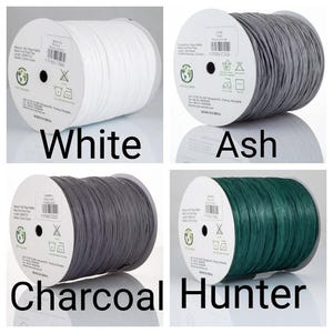 May include: Four spools of raffia yarn in white, ash, charcoal, and hunter green. Each spool features a central label with product details, including material, length, and width, alongside care icons and a recycling symbol. The yarn is wound tightly.