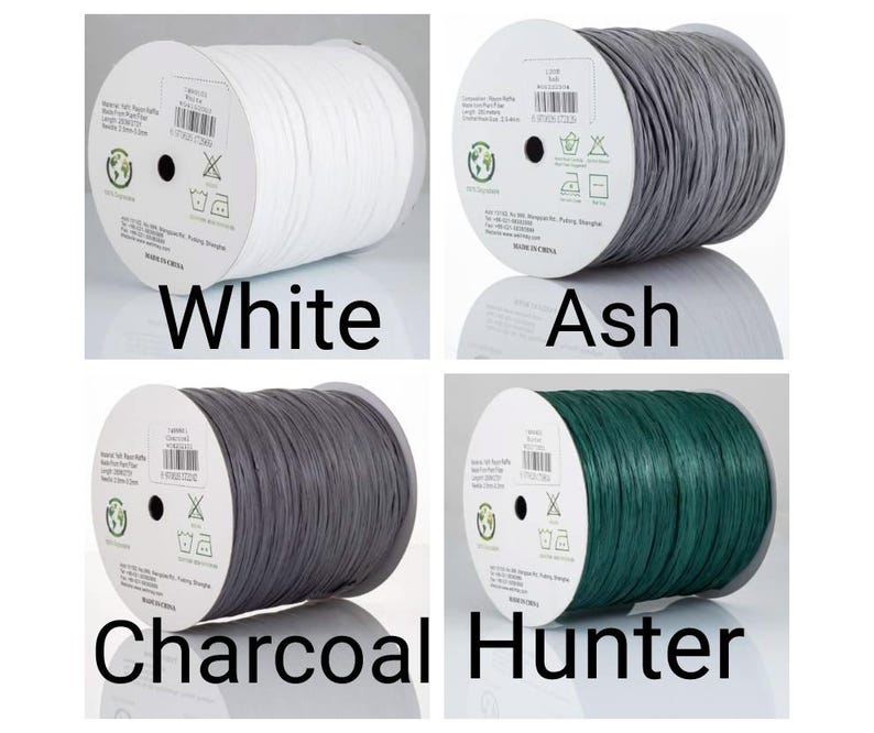 May include: Four spools of raffia yarn in white, ash, charcoal, and hunter green. Each spool features a central label with product details, including material, length, and width, alongside care icons and a recycling symbol. The yarn is wound tightly.