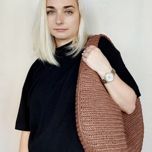 May include: A large, crescent-shaped hobo bag crafted from a textured, woven brown material. The bag features a continuous, integrated shoulder strap and a dense, braided pattern throughout its surface, providing a structured yet flexible form.