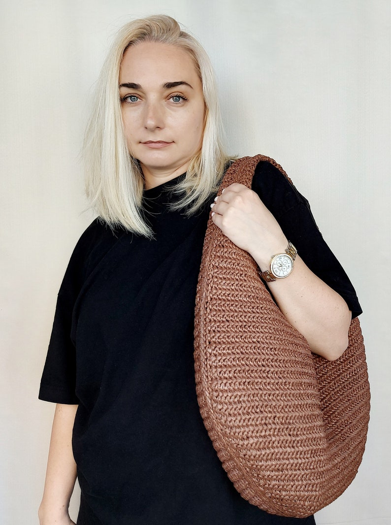 May include: A large, crescent-shaped hobo bag crafted from a textured, woven brown material. The bag features a continuous, integrated shoulder strap and a dense, braided pattern throughout its surface, providing a structured yet flexible form.