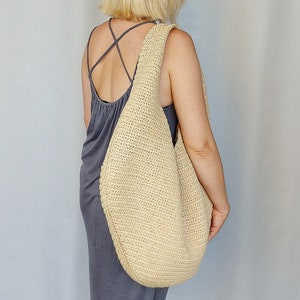 May include: A large, beige, crescent-shaped hobo bag crafted from a textured, woven material. The bag features a wide, integrated shoulder strap and a slouchy, unstructured silhouette that hangs against the side of the body.