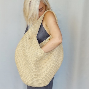 May include: A large, beige, crescent-shaped hobo bag crafted from a textured, woven material. The bag features a wide, integrated shoulder strap and a slouchy, unstructured silhouette with a visible stitch pattern throughout the exterior surface.