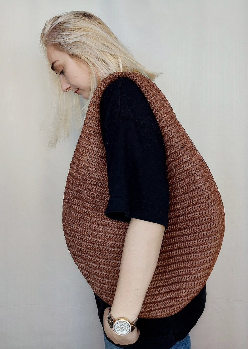 May include: A large, crescent-shaped hobo bag crafted from a brown, textured, woven material. The bag features a wide, integrated shoulder strap and a continuous, ribbed pattern throughout its surface, creating a rounded, voluminous silhouette.