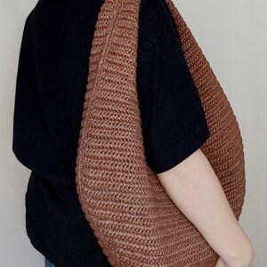 May include: A large, crescent-shaped hobo bag crafted from a brown, woven material with a distinct ribbed texture. The bag features a wide, integrated shoulder strap and a slouchy, unstructured silhouette that hangs against a black t-shirt and blue jeans.