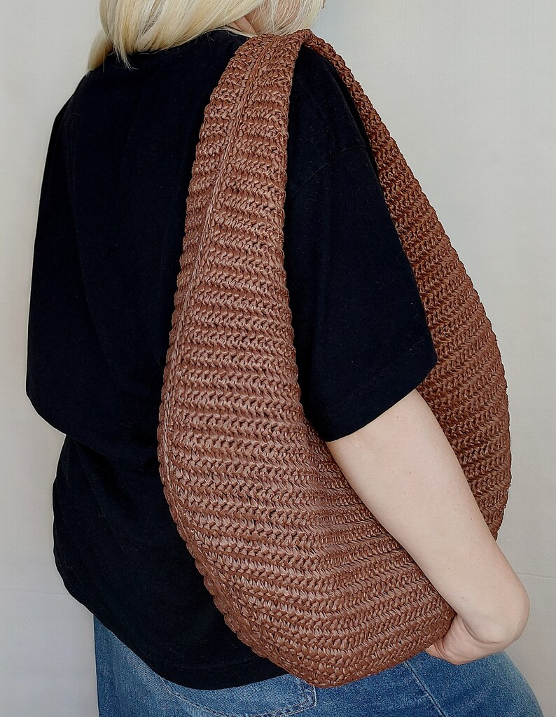 May include: A large, crescent-shaped hobo bag crafted from a brown, woven material with a distinct ribbed texture. The bag features a wide, integrated shoulder strap and a slouchy, unstructured silhouette that hangs against a black t-shirt and blue jeans.