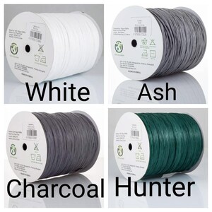 May include: Four spools of raffia yarn in white, ash, charcoal, and hunter green colors. Each spool features a central label with product details, including material composition, length, and care instructions, with a circular hole in the center.