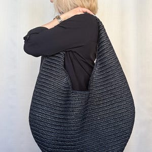 May include: A large, slouchy hobo-style shoulder bag crafted from dark charcoal gray woven material. The bag features a continuous, integrated strap design that forms a deep, rounded crescent shape with a textured, braided surface pattern.