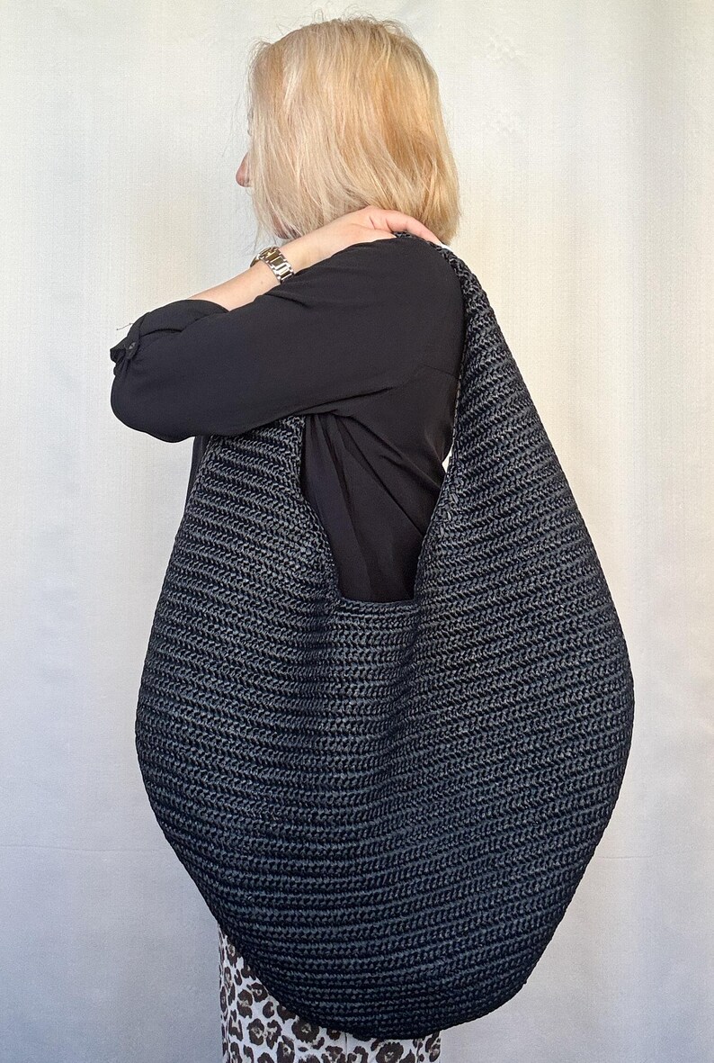 May include: A large, slouchy hobo-style shoulder bag crafted from dark charcoal gray woven material. The bag features a continuous, integrated strap design that forms a deep, rounded crescent shape with a textured, braided surface pattern.