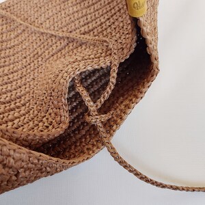 May include: A light brown woven bag featuring a textured, crocheted surface with a braided drawstring closure. A small, yellow leather-like tab with embossed text is attached to the upper side, providing a subtle contrast to the tan material.