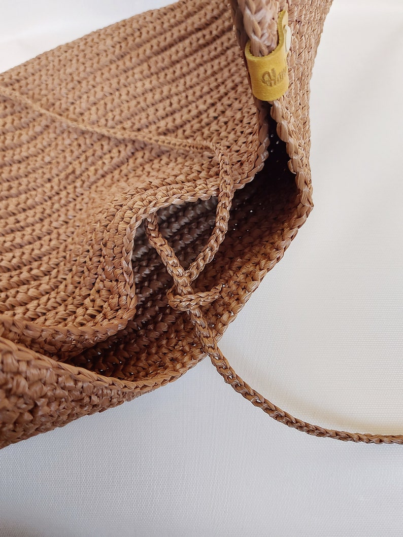 May include: A light brown woven bag featuring a textured, crocheted surface with a braided drawstring closure. A small, yellow leather-like tab with embossed text is attached to the upper side, providing a subtle contrast to the tan material.