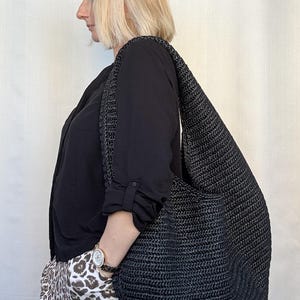 May include: A large, black hobo-style shoulder bag crafted from a woven, textured material. The bag features a wide, integrated strap that rests comfortably over the shoulder, with a slouchy, unstructured silhouette and a deep, open main compartment.