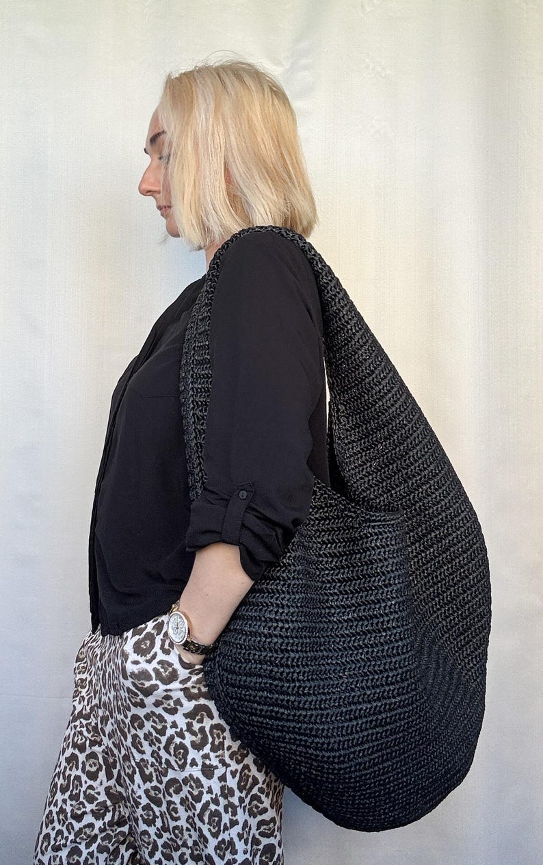 May include: A large, black hobo-style shoulder bag crafted from a woven, textured material. The bag features a wide, integrated strap that rests comfortably over the shoulder, with a slouchy, unstructured silhouette and a deep, open main compartment.