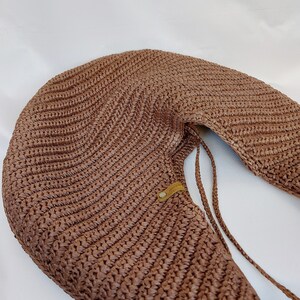 May include: A crescent-shaped brown woven shoulder bag featuring a textured, ribbed pattern throughout the body. The bag includes a small, rectangular tan leather-like tag with a metallic rivet and thin braided straps attached to the side.