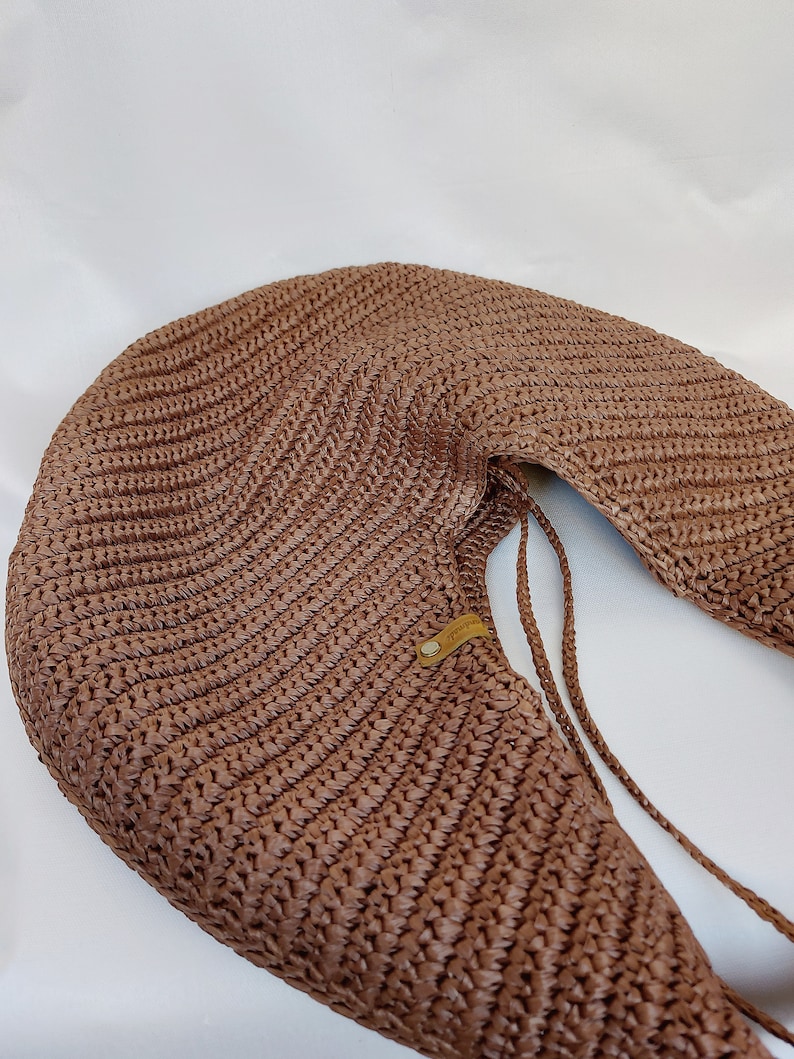 May include: A crescent-shaped brown woven shoulder bag featuring a textured, ribbed pattern throughout the body. The bag includes a small, rectangular tan leather-like tag with a metallic rivet and thin braided straps attached to the side.