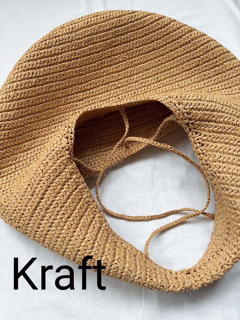 May include: A tan, round, woven hobo bag with a textured surface and thin, matching woven straps. The bag features a wide, open top and a consistent, dense braided pattern throughout its construction. The word Kraft is printed in black text.