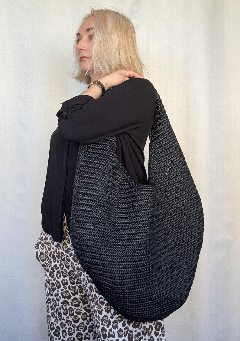 May include: A large, dark gray and black woven shoulder bag. The bag has a wide strap and a rounded shape. The bag is made of a textured material and appears to be handmade. The bag is worn over the shoulder.